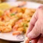 Oven-roasted shrimp on skewers that have been coated in garlic butter with one shrimp being dipped in garlic butter.