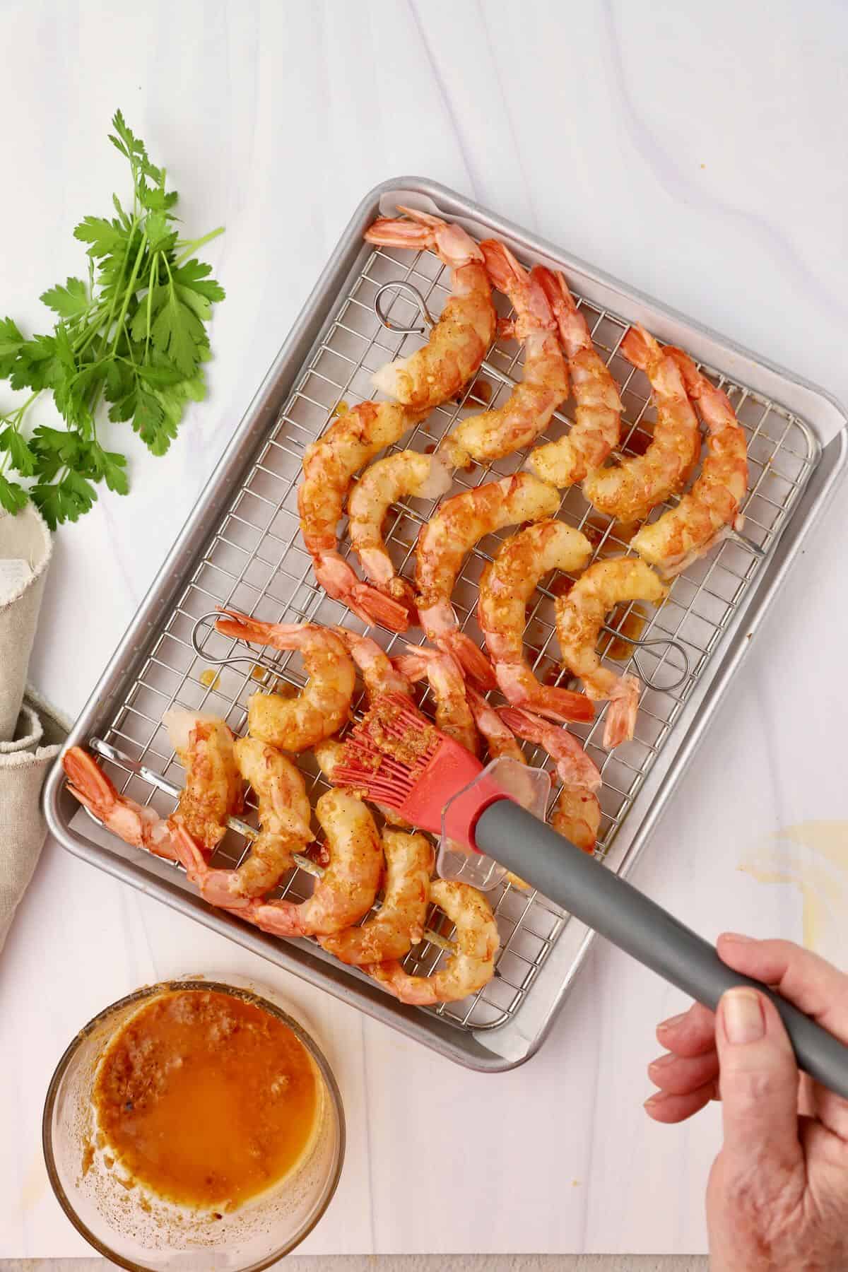 Royal red shrimp on skewers on a metal baking rack with garlic butter being brushed on.
