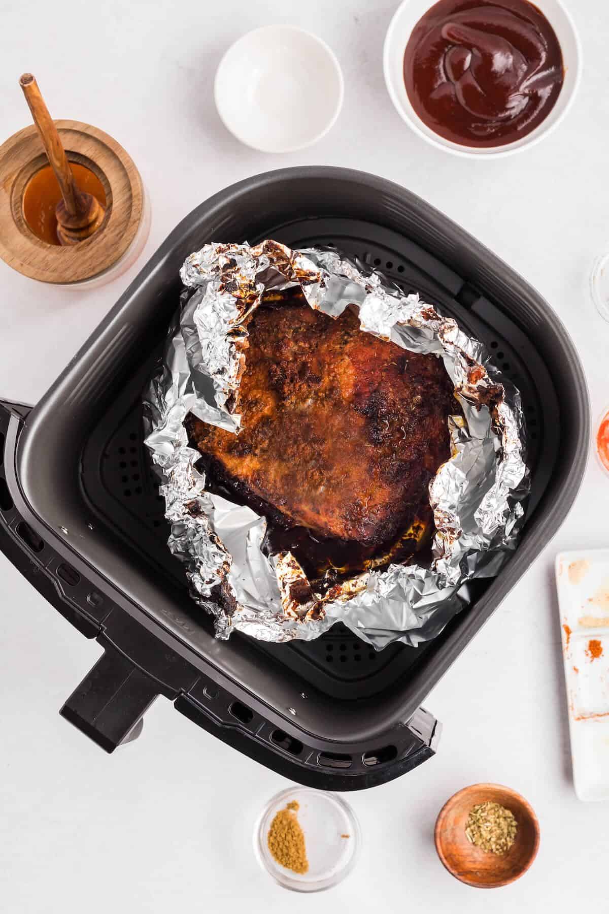 A pork butt in an air fryer basket wrapped in foil is being flipped over.