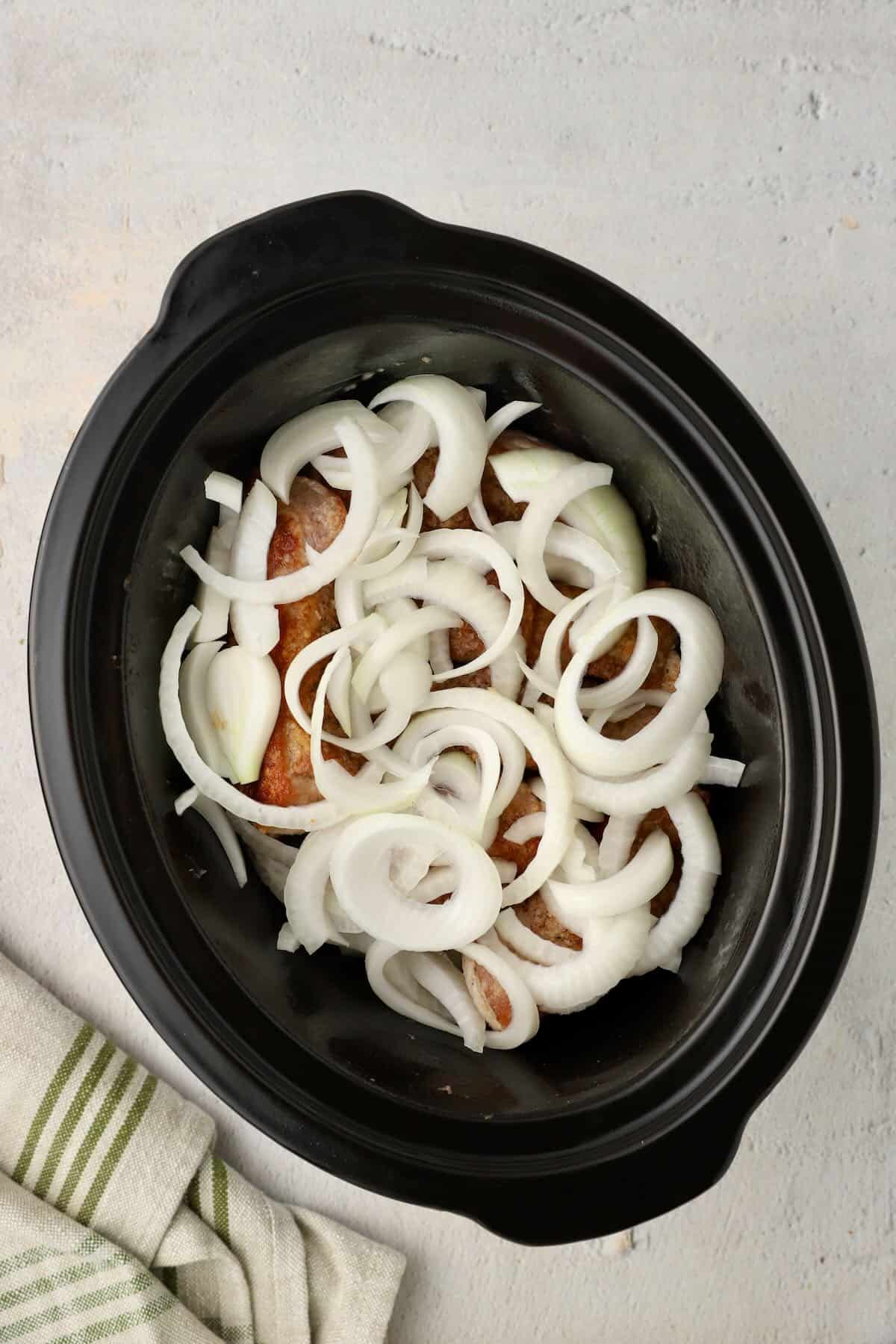 A black slow cooker bowl full of sliced onions and boneless pork ribs.
