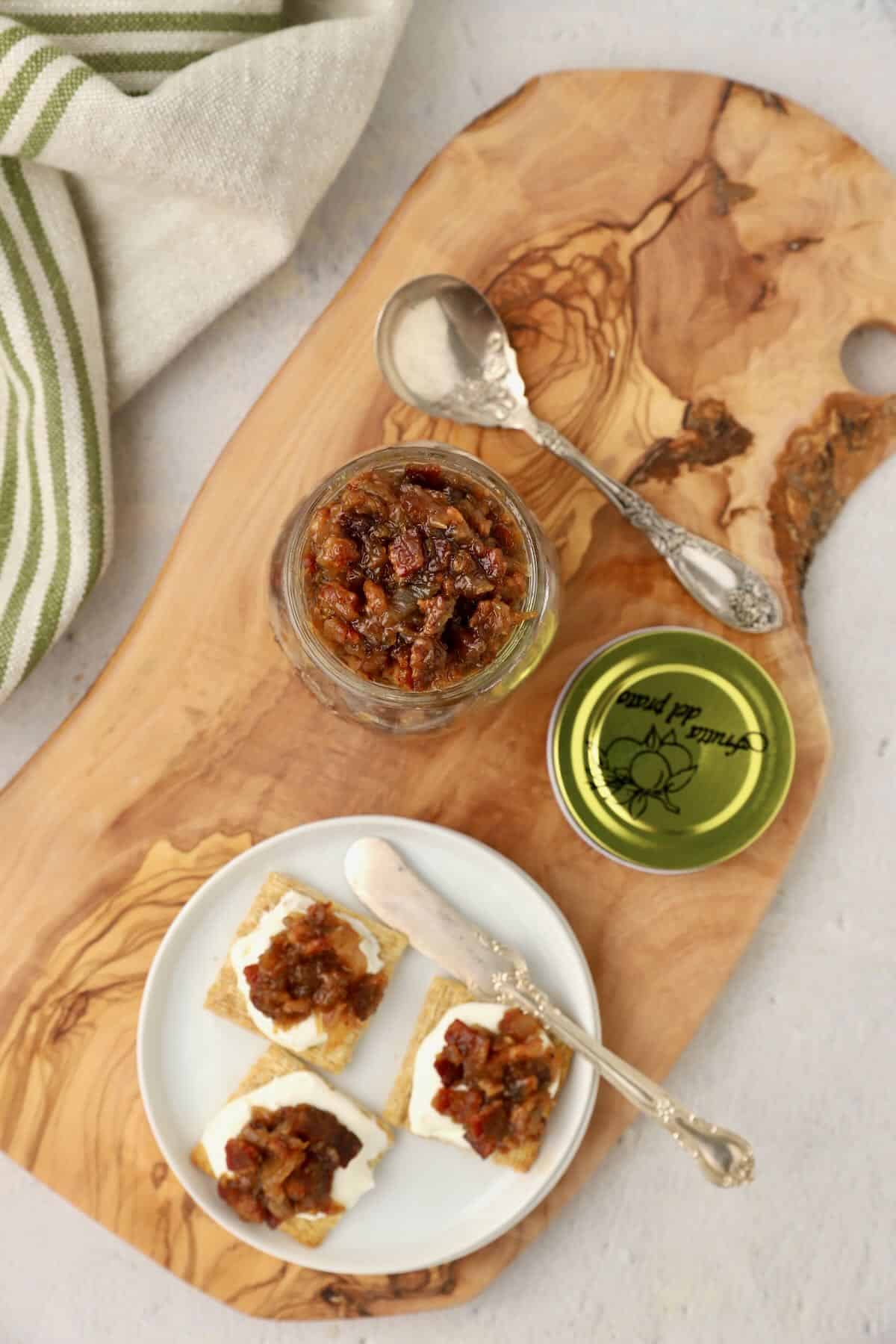 A jar of bourbon bacon onion jam on a cutting board with crostini topped with cream cheese and jam.