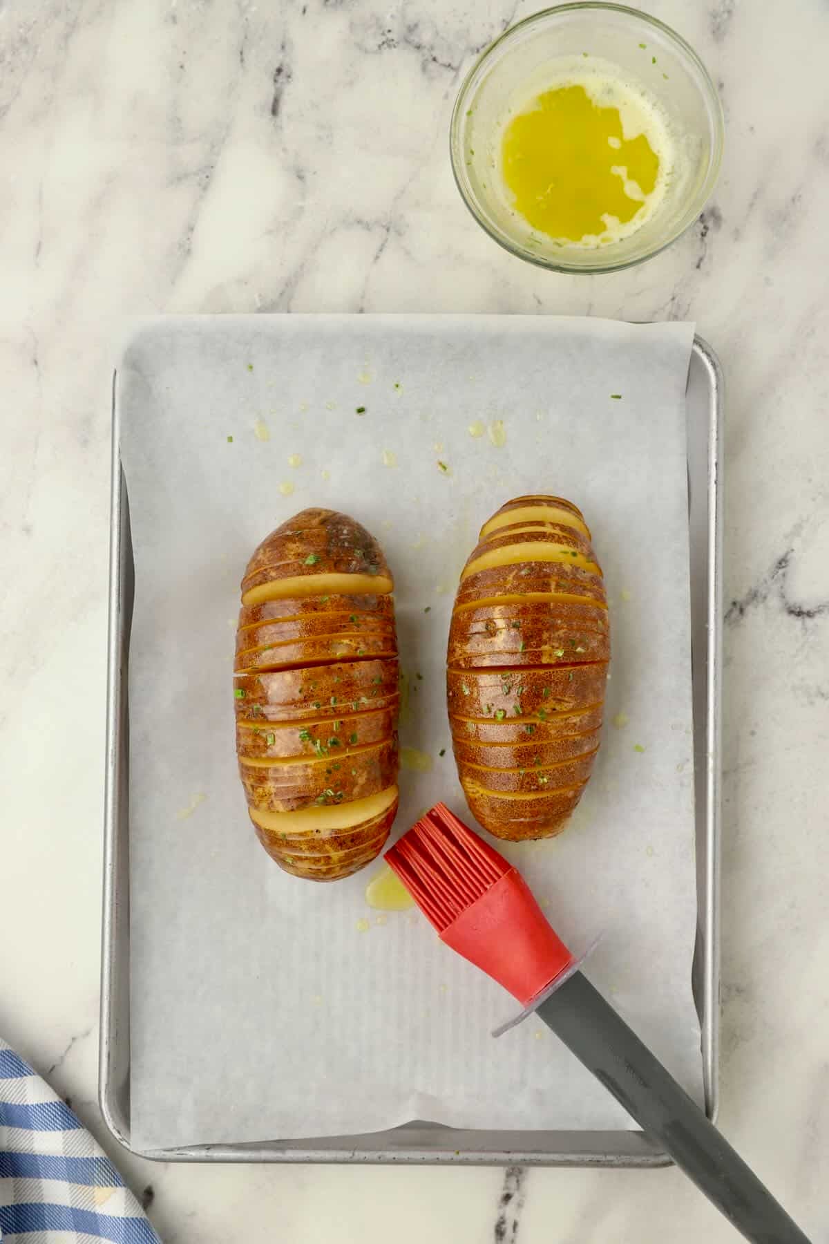 Two hasselback potatoes with slices cut and basted with butter on a baking sheet, ready for the oven.