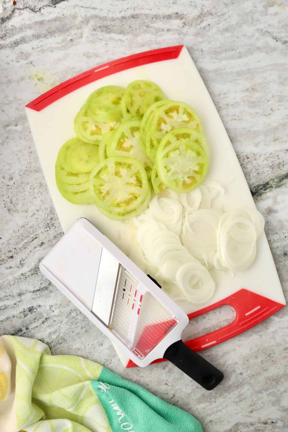 Thinly slices green tomatoes and onions on a cutting board with a mandoline.