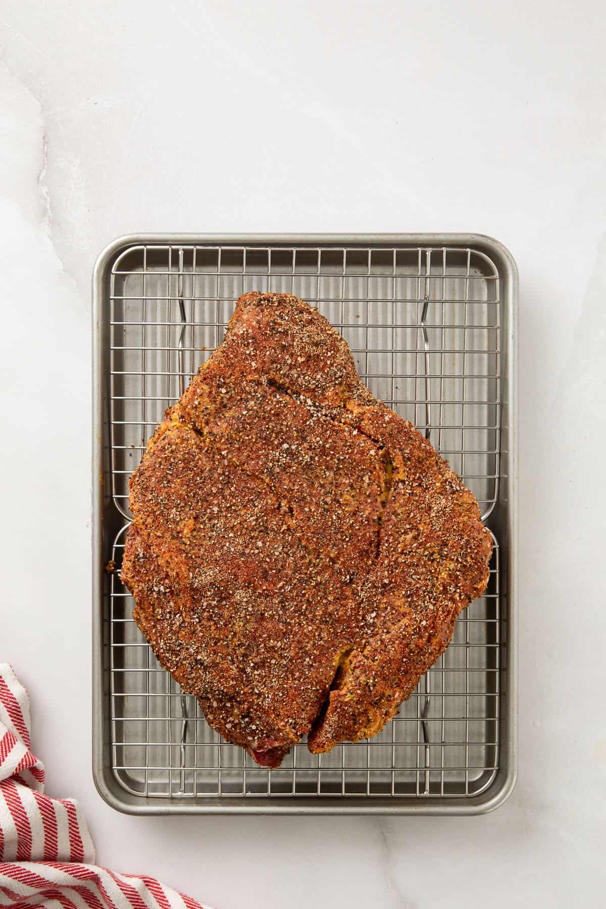 A raw chuck roast coated with a dry brine rub on a wire grate over a rimmed baking sheet.