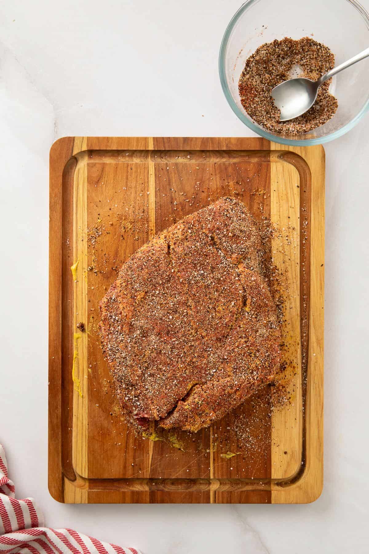 A chuck roast on a cutting board coated with a mustard binder and dry brine rub.