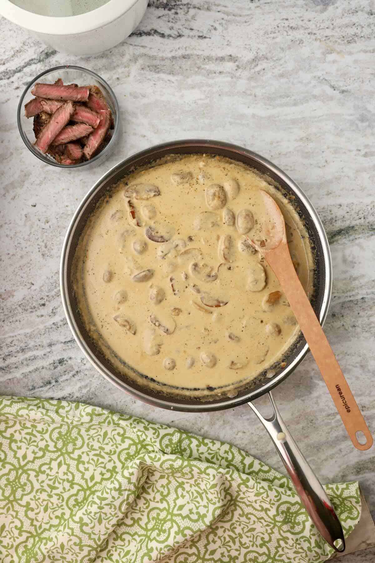 A skillet with cooked mushrooms and onions, with beef broth and heavy cream added.