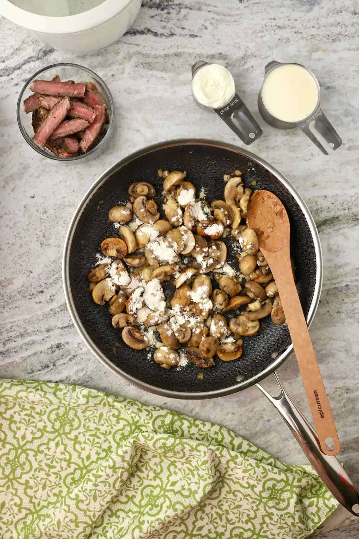 Sliced mushrooms and chopped onions are cooking in a skillet with flour sprinkled over the top.