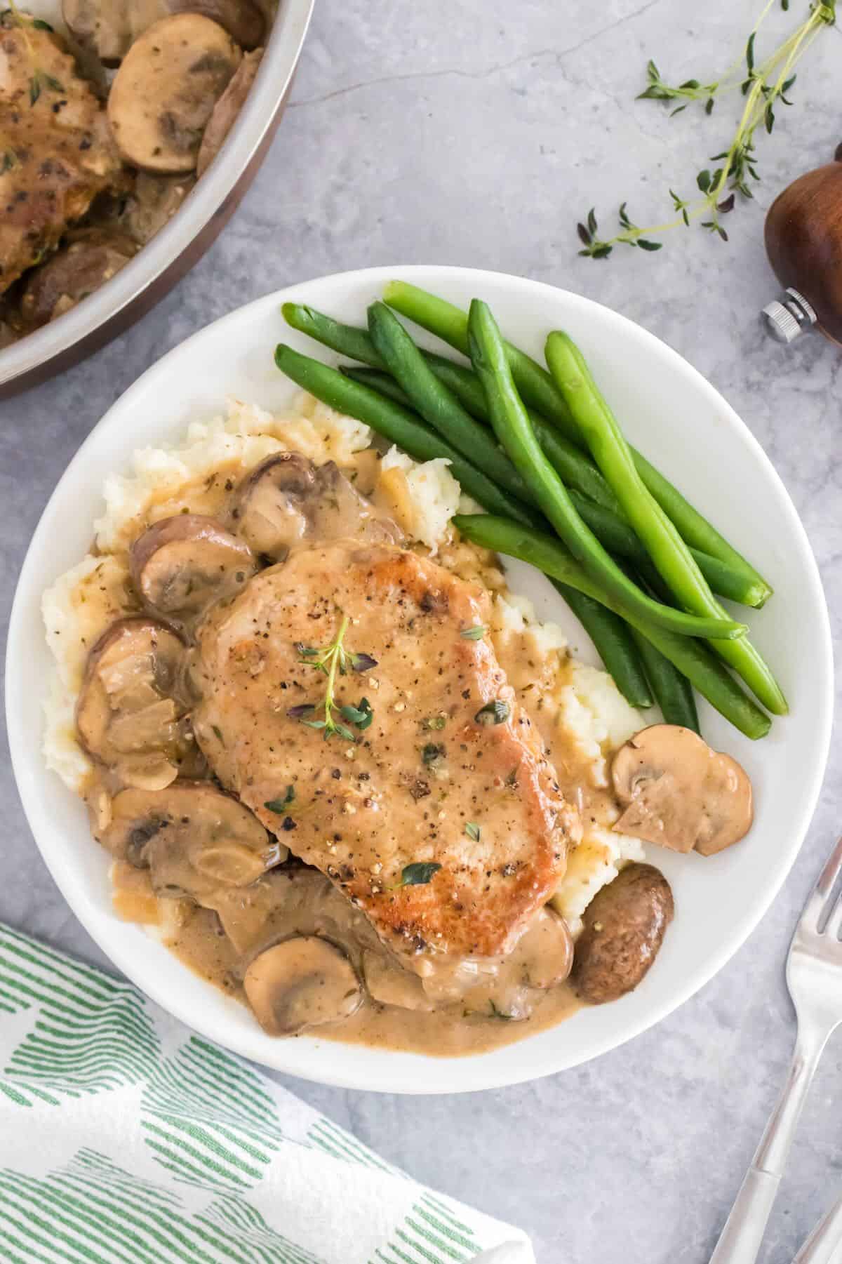 A pan seared pork chop topped with a mushroom pan sauce on a plate with green beans.