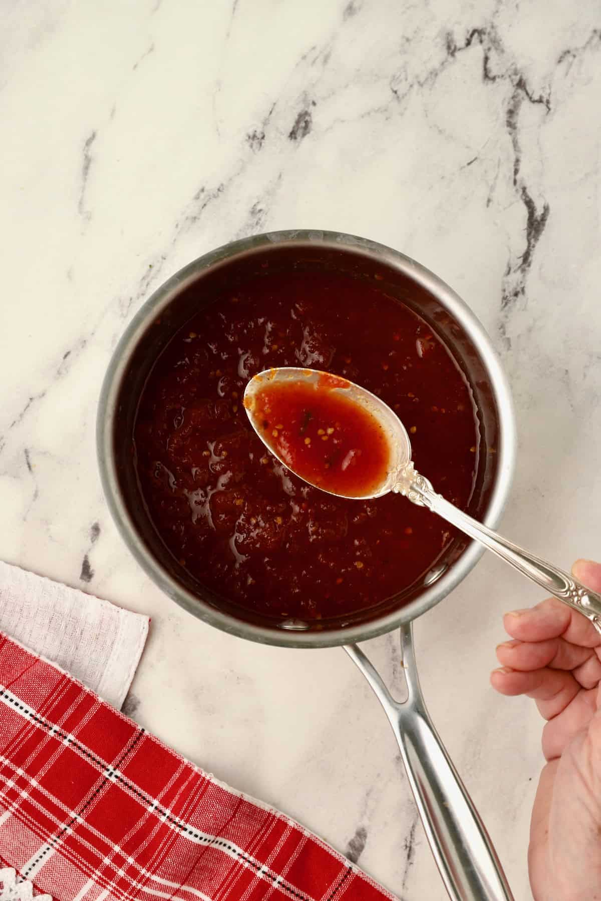 Tomato jam is simmering in a saucepan on the stove with a spoon.