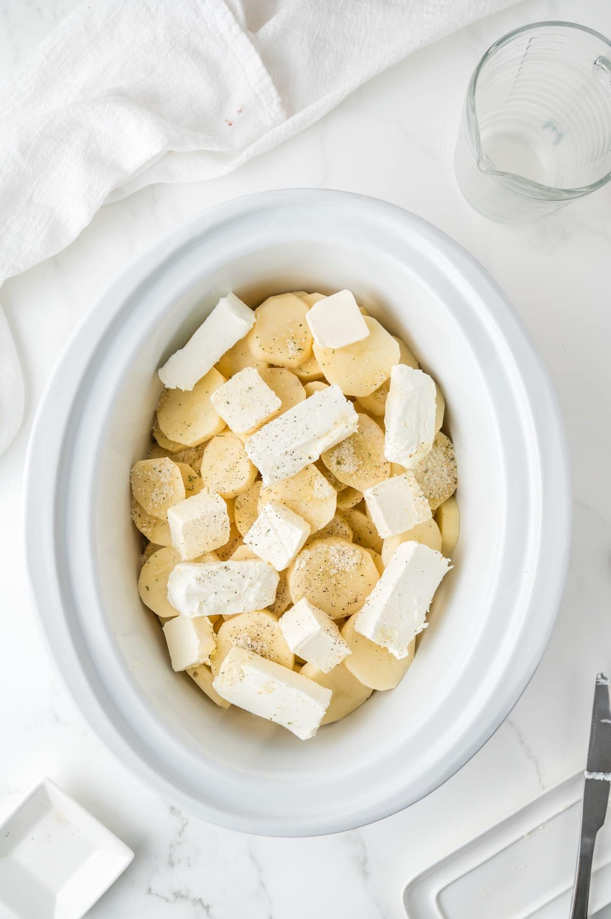 A white crock pot bowl full of sliced potatoes topped with pats of butter and cubes of cream cheese.