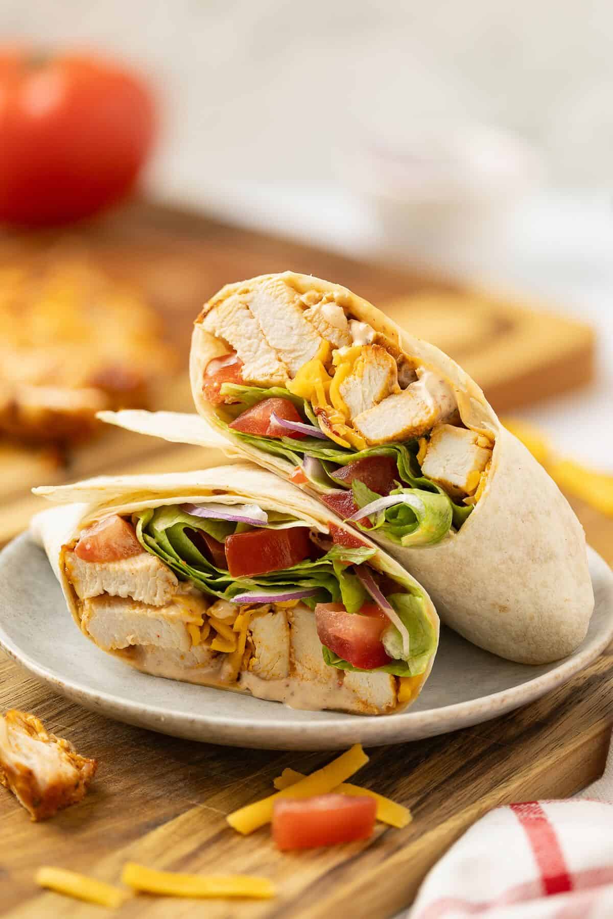 A grilled chicken wrap full of grilled chicken, lettuce, tomato, and shredded cheese, cut in half on a plate on a cutting board.